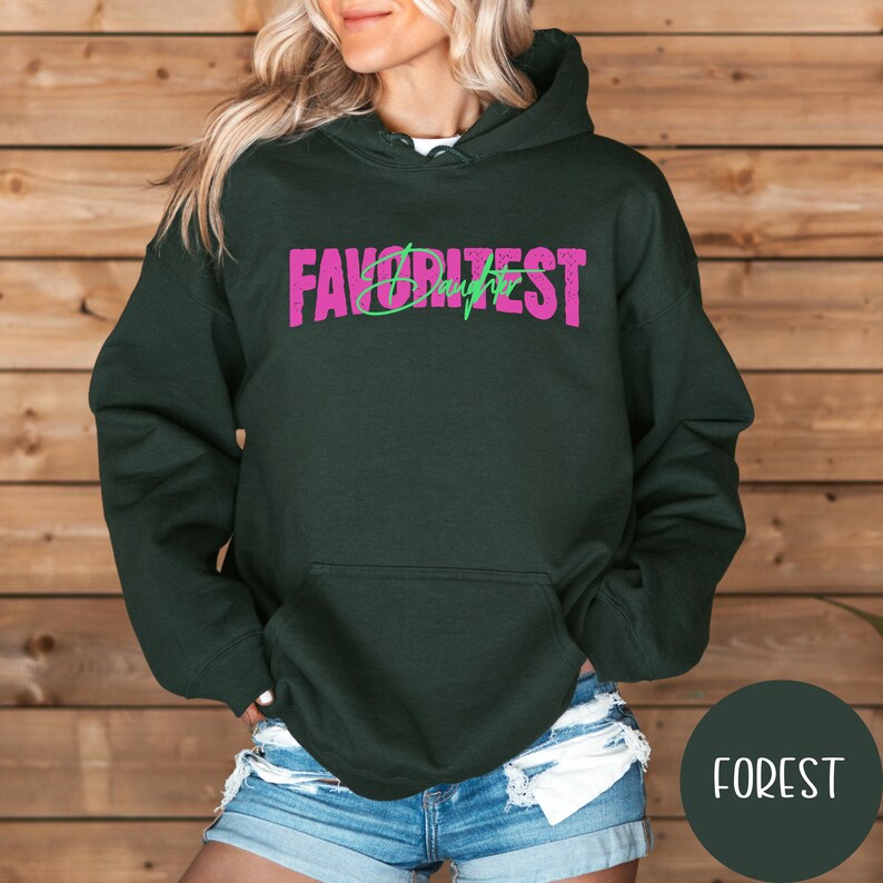 May include: A dark green hoodie with the text "FAVORITEST Daughter" in pink and green. The sweatshirt features a front pocket and a drawstring hood. The background is a wooden wall.