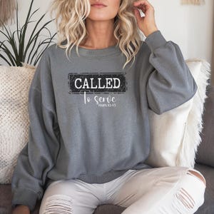 Könnte beinhalten: Ein graues Sweatshirt mit dem Text "CALLED To serve mark 10:45" in einem schwarz-weißen rechteckigen Design. Das Sweatshirt hat lange Ärmel und einen Rundhalsausschnitt. Das Model trägt eine weiße Hose.