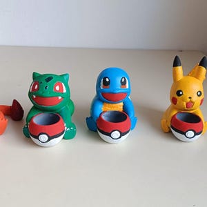 Puede incluir: Cuatro pequeñas figuras pintadas a mano de criaturas de dibujos animados, incluyendo un lagarto naranja, una criatura verde, una tortuga azul y un ratón amarillo, cada una sosteniendo un pequeño recipiente redondo rojo, blanco y negro.