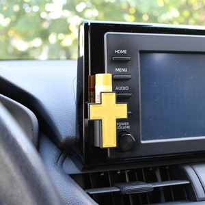 May include: A gold cross car air freshener hanging from the air vent of a car. The cross is attached to a black plastic holder. The car's dashboard is visible in the background.
