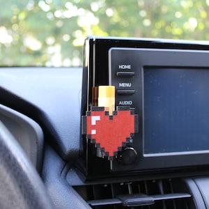 May include: A red pixelated heart-shaped car air freshener hanging from the air vent of a car dashboard. The air freshener is next to a car radio with buttons labeled "Home", "Menu", and "Audio".