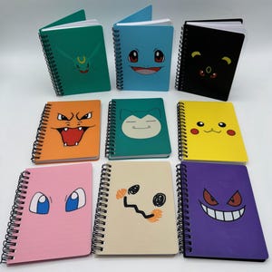 May include: A collection of spiral-bound notebooks with colorful covers featuring various cartoon character faces. The notebooks are in shades of green, blue, black, orange, teal, yellow, pink, beige, and purple. The notebooks are blank inside.