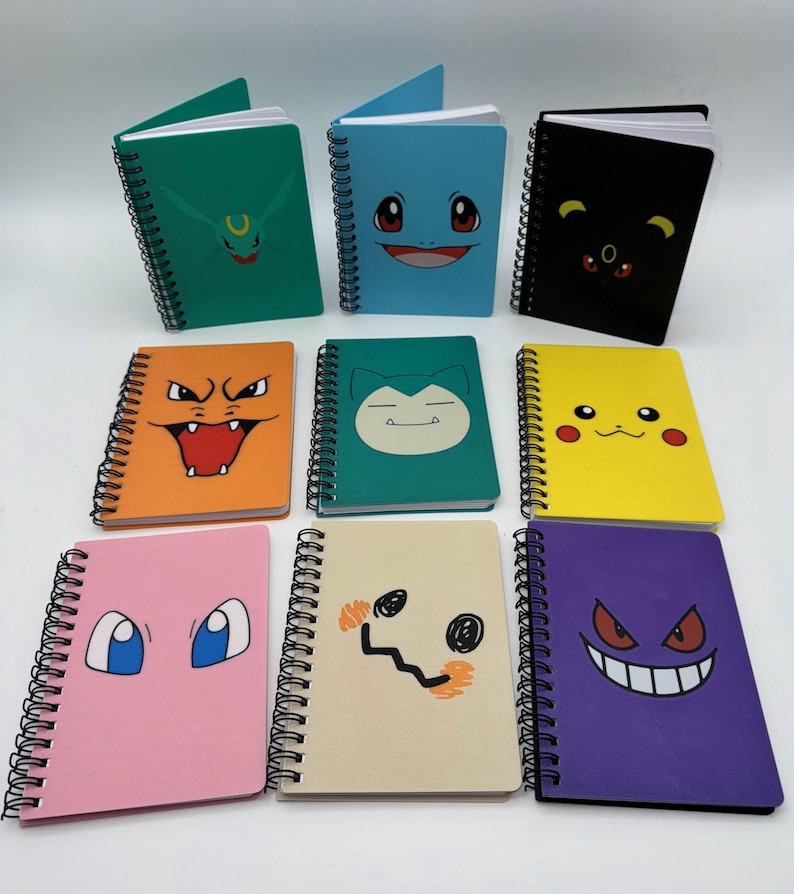 May include: A collection of spiral-bound notebooks with colorful covers featuring various cartoon character faces. The notebooks are in shades of green, blue, black, orange, teal, yellow, pink, beige, and purple. The notebooks are blank inside.