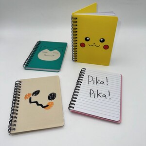 May include: Four spiral-bound notebooks with character designs. A yellow notebook features a Pikachu face, a green notebook has a Snorlax face, and a tan notebook has a Mimikyu face. A fourth notebook has the text "Pika! Pika!" on the lined pages.