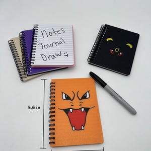 May include: A collection of spiral-bound notebooks. One orange notebook features a cartoon face, measuring 4 inches wide and 5.6 inches tall. Other notebooks are black, purple, and tan. One notebook has the words "Notes Journal Draw" written on the cover.