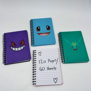 May include: Four spiral-bound notebooks with colorful covers. One purple notebook features a smiling face, a blue notebook has a smiling face, and a green notebook has a face. A white notebook has the text "120 Pages / 60 sheets".