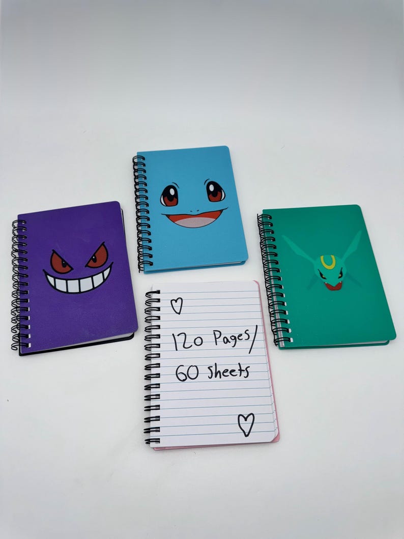 May include: Four spiral-bound notebooks with colorful covers. One purple notebook features a smiling face, a blue notebook has a smiling face, and a green notebook has a face. A white notebook has the text "120 Pages / 60 sheets".