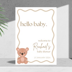 May include: A white baby shower invitation with a brown teddy bear illustration. The text reads "hello baby." and "welcome to Rachael's baby shower 21 JANUARY 2024".
