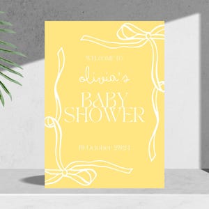 May include: A yellow baby shower invitation with white text and a white ribbon design. The text reads "Welcome to Olivia's Baby Shower" and "19 October 2024".