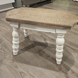 May include: A small wooden stool featuring a rectangular top with a light brown, textured finish and four white, turned legs. The stool has a distressed appearance with visible paint wear on the legs and edges of the wooden top surface.