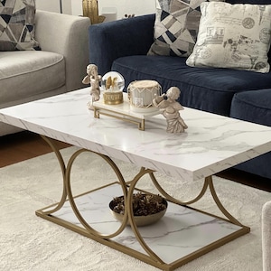 May include: A rectangular coffee table featuring a white faux marble top and a lower shelf, supported by a gold-toned metal frame with circular side accents. The table holds a small decorative tray with a snow globe and a candle.