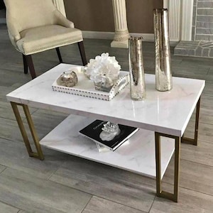 May include: A rectangular coffee table with a white faux marble top and a lower shelf, supported by four gold-toned metal legs. The table holds a white tray with a floral arrangement, two tall metallic vases, and a black book on the lower shelf.