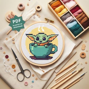 Puede incluir: Un patrón de punto de cruz con un personaje verde en una taza de té turquesa, sobre un fondo amarillo. El diseño está enmarcado en un aro de madera, rodeado de suministros de bordado, incluyendo hilo, tijeras y agujas. El texto "PDF to print or download" es visible.