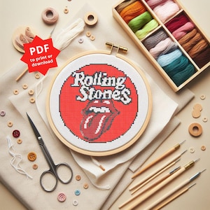 Puede incluir: Un bastidor de bordado de punto de cruz con el logotipo de los Rolling Stones, un círculo rojo con el nombre de la banda y el gráfico de la lengua. La imagen incluye suministros de bordado como hilo, tijeras y herramientas de madera.