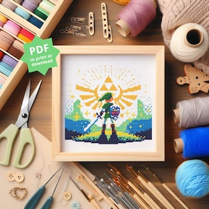 Op de afbeelding: Een ingelijste kruissteek met een pixelontwerp van een videogamepersonage met een zwaard en schild, tegen een achtergrond van een gouden embleem en een blauw en groen landschap. De lijst is van lichtgekleurd hout. Tekst op de afbeelding zegt "PDF to print or download."