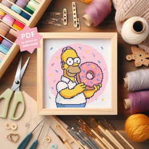 Peut inclure: Motif de point de croix représentant Homer Simpson tenant un donut rose. L'œuvre est encadrée dans un cadre en bois clair. Des fournitures de bricolage, dont des ciseaux, du fil et des aiguilles, entourent l'image. Le texte sur l'image indique "PDF to print or download".