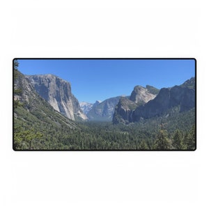 May include: A landscape image showing a valley with high rock formations and dense green trees under a clear blue sky. The scene presents a panoramic view of a natural environment.