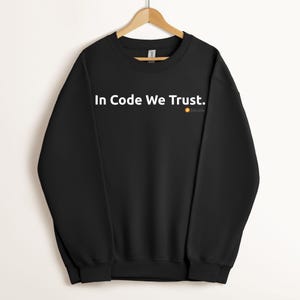 May include: A black long-sleeve crewneck sweatshirt featuring the white text In Code We Trust. printed across the chest. Below the text is a small orange circular Bitcoin logo. The garment is displayed on a wooden hanger against a plain background.