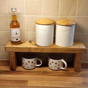 May include: A wooden shelf displays a bottle, two white canisters labeled "tea" and "sugar", and two mugs with colorful polka dots. The shelf is set against a tiled wall and a wooden countertop.