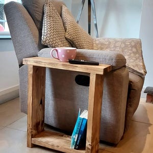 May include: A wooden side table with a shelf, featuring a light brown finish. The table is next to a grey armchair with cushions and a pink mug on top of the table. Three books are on the shelf.