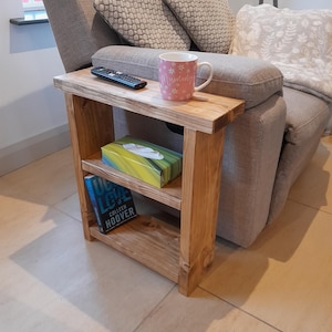 May include: A wooden side table with two shelves, a book, a box of tissues, and a pink mug with the word "morning" on it. The table is next to a grey armchair with a white throw blanket.