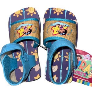 May include: Pair of blue and purple sandals featuring Dora the Explorer. The sandals have a purple base with a pattern of suns and bamboo. The straps are blue with a woven design and a Dora the Explorer graphic. A tag with the Dora the Explorer logo is attached.