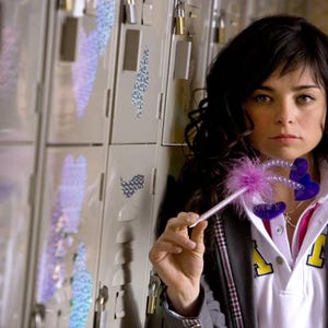 May include: A woman in a white collared shirt and gray blazer holds a pink and purple butterfly wand. The background features lockers with iridescent stickers. The woman has dark hair and is looking at the camera.