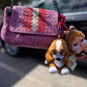 May include: A purple leather handbag with a woven design and a chain strap. The flap features a red and white pattern. Attached are two plush keychains: a sad-eyed dog and a cartoon character.
