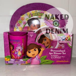 May include: A Dora the Explorer personalized 3-piece dinner set in a pink box. The set includes a plate, bowl, and tumbler. The box features Dora and other characters, with the Nickelodeon logo.