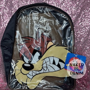 May include: Black and clear backpack featuring a Taz cartoon graphic. The backpack has a black zipper and straps. The Taz character is depicted with a yellow face, red hair, and a wide grin. A tag with the text "LO NAKED DENIM" is attached.