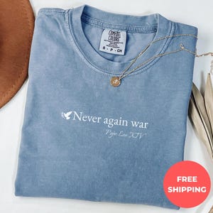 May include: A blue Comfort Colors t-shirt with the text "Never again war" and "Pope Leo XIV" printed in white. A gold necklace with a pendant is draped across the shirt. A brown hat is visible in the upper left corner.