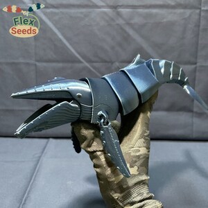 Puede incluir: Una escultura de ballena articulada impresa en 3D de color azul oscuro. La ballena est&aacute; posada con la boca abierta y la cola levantada. La escultura est&aacute; hecha de m&uacute;ltiples piezas segmentadas que permiten la articulaci&oacute;n.