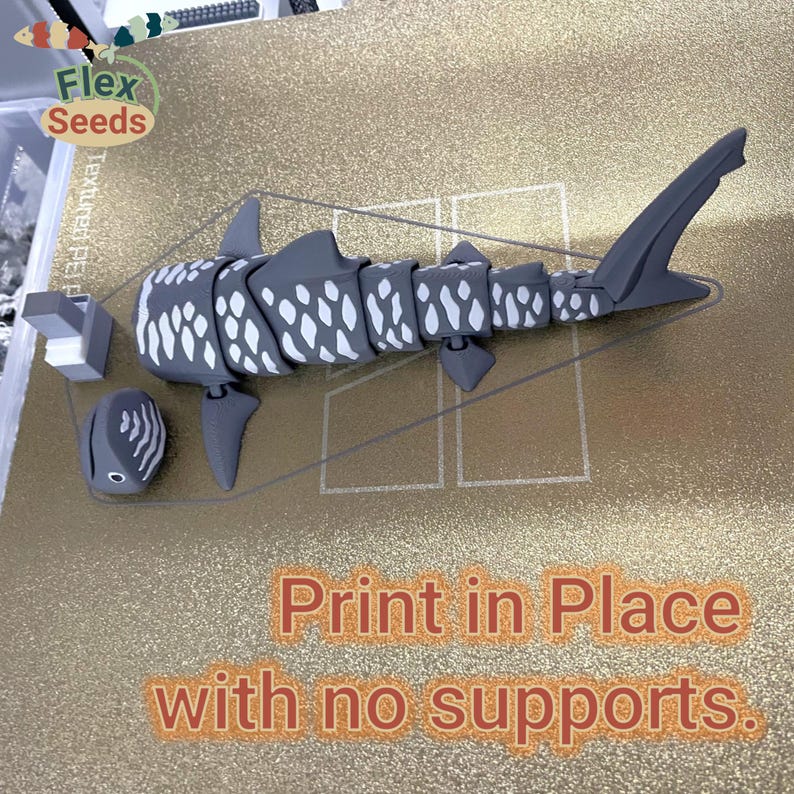 May include: A 3D printed model of a whale shark, printed in place with no supports. The model is gray with white spots and is printed on a textured gold surface. The text "Print in Place with no supports." is printed below the model.