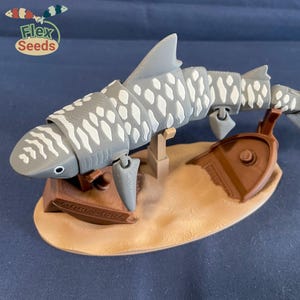 May include: A gray and white articulated shark toy with a white and brown shipwreck base. The shark is posed as if it is attacking the shipwreck. The toy is made by Flex Seeds.