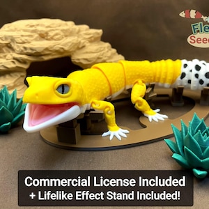 Puede incluir: Una figura articulada de gecko leopardo amarilla y blanca con manchas negras en la cola, con la boca abierta y el interior rosa, colocada sobre un soporte ovalado marrón con una formación rocosa y dos plantas suculentas verdes.