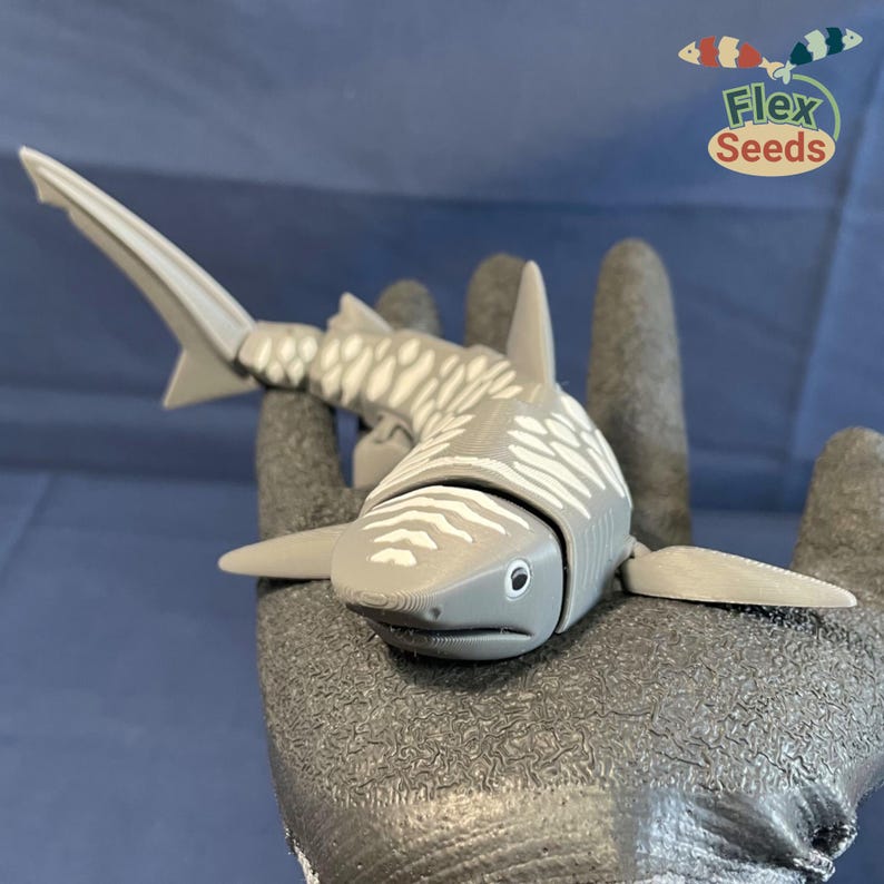 May include: A gray and white plastic toy shark with a segmented body. The shark has a long, pointed snout and a dorsal fin. The toy is posed on a gray surface.