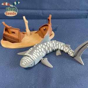 May include: A gray and white tiger shark toy with articulated joints, swimming near a brown toy shipwreck with the text "Tiger Shark" on the side. The shipwreck is partially submerged in a tan sand-colored base.