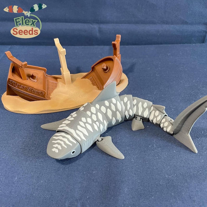 May include: A gray and white tiger shark toy with articulated joints, swimming near a brown toy shipwreck with the text "Tiger Shark" on the side. The shipwreck is partially submerged in a tan sand-colored base.