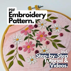 May include: A PDF embroidery pattern featuring a floral design on light pink fabric held in a yellow hoop. The embroidery includes white daisies, pink flowers, green leaves, and small dark red knots. Text reads: PDF Embroidery Pattern. incl. Step-by-Step Tutorial & Videos.