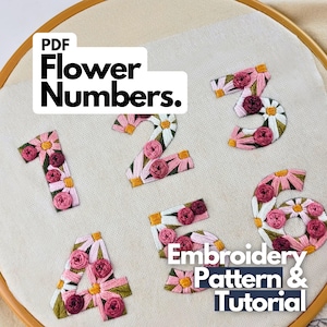 Op de afbeelding: Geborduurde cijfers 1 tot en met 6, elk versierd met roze en witte bloemmotieven, worden getoond op crèmekleurige stof in een houten borduurring. De tekst "PDF Flower Numbers" en "Embroidery Pattern & Tutorial" zijn ook zichtbaar, voor een borduurpatroon.