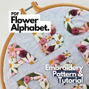 Op de afbeelding: Een borduurring houdt een lichtgrijze stof vast met geborduurde bloemenalfabetletters. De letters zijn versierd met roze, witte en gele bloemen en donkerrode rozenaccenten. De tekst "PDF Flower Alphabet" en "Embroidery Pattern & Tutorial" is ook zichtbaar.