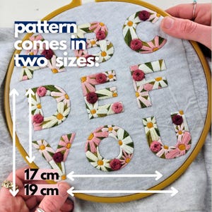 K&ouml;nnte beinhalten: Gestickte Buchstaben A bis V, jeweils mit G&auml;nsebl&uuml;mchen- und Rosenmotiven verziert, werden in einem h&ouml;lzernen Stickrahmen gezeigt. Der Text "pattern comes in two sizes" ist sichtbar. Die Stickerei misst 17 cm und 19 cm.