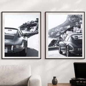 Op de afbeelding: Twee ingelijste zwart-wit prints van klassieke auto's op een kustweg. De auto's zijn donkergrijs, met de oceaan en een heuvelstad op de achtergrond. De lijsten zijn zwart en de prints worden op een witte muur getoond.