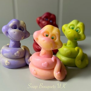 May include: Four handmade soap sculptures of snakes in different colours: purple, pink, green, and red. Each snake is curled up with its tail in its mouth and has a heart pattern on its body. The soaps are labelled "Soap Bouquets UK".