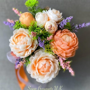 May include: A bouquet of peach and white soap flowers with green leaves and purple lavender accents. The bouquet is arranged in a white container and has the text "Soap Bouquets UK" at the bottom.