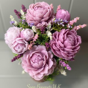 May include: A bouquet of purple soap flowers with green leaves and small pink flowers. The bouquet is arranged in a vase and the text "Soap Bouquets UK" is visible at the bottom of the image.