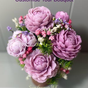 May include: A bouquet of purple and pink soap flowers arranged in a brown pot. The flowers are made to look like peonies and are surrounded by small green leaves and white and pink flowers. The text "Soap Bouquets UK" is at the bottom of the image.