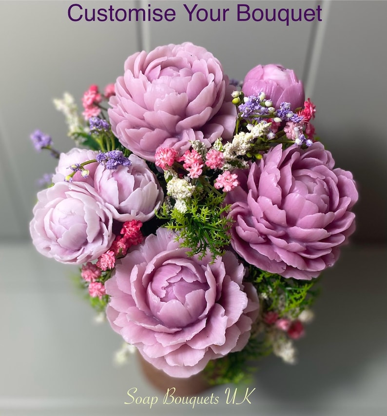 May include: A bouquet of purple and pink soap flowers arranged in a brown pot. The flowers are made to look like peonies and are surrounded by small green leaves and white and pink flowers. The text "Soap Bouquets UK" is at the bottom of the image.