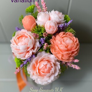 May include: A bouquet of peach and white soap flowers with green leaves and purple accents. The flowers are arranged in a vase and the bouquet is labeled "Soap Bouquets UK".