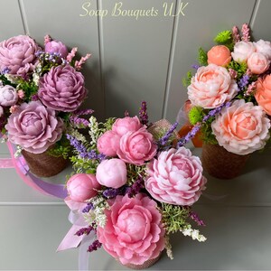 May include: Three soap flower bouquets in different shades of pink and peach. Each bouquet is arranged in a small brown basket and features a variety of flowers, including peonies, lavender, and greenery. The bouquets are arranged on a gray background.